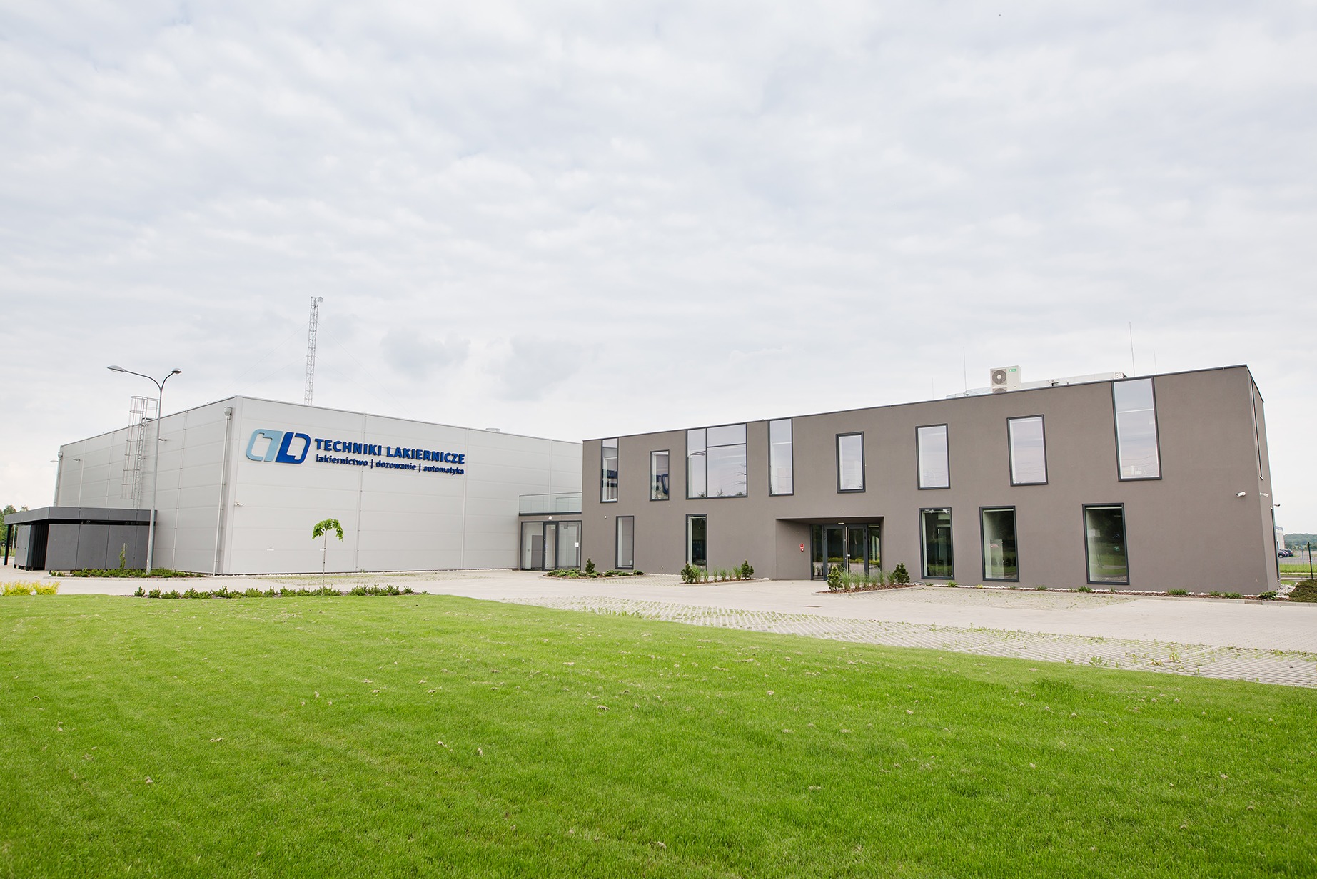 Nowoczesny budynek przemysłowy z logo firmy 'Techniki Lakiernicze' na elewacji, otoczony trawnikiem i kostką brukową.