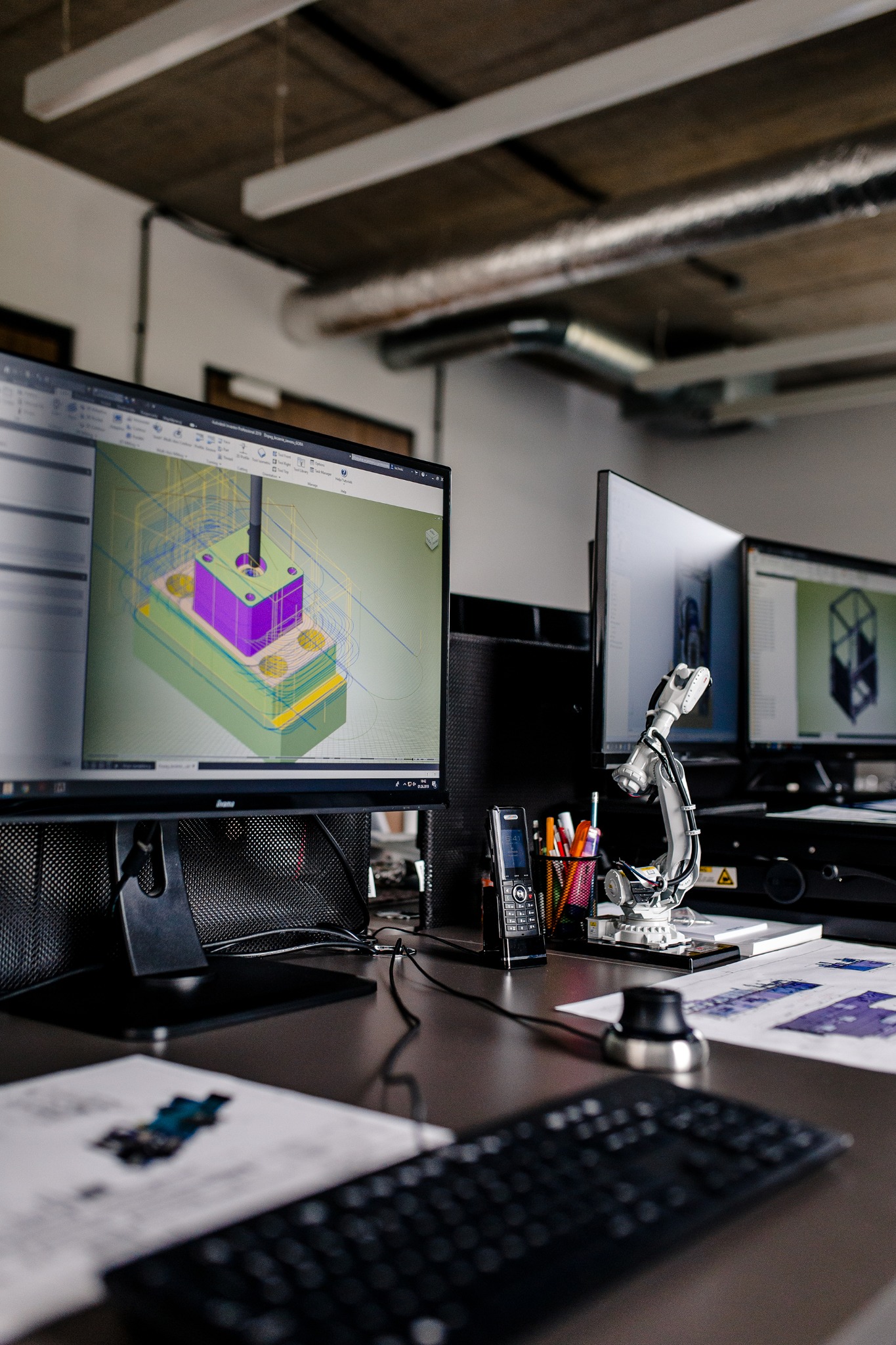 Stanowisko projektowe z trzema monitorami, na pierwszym wizualizacja 3D obrabianego elementu, obok model robota przemysłowego, telefon stacjonarny i klawiatura, na biurku leżą wydruki techniczne.