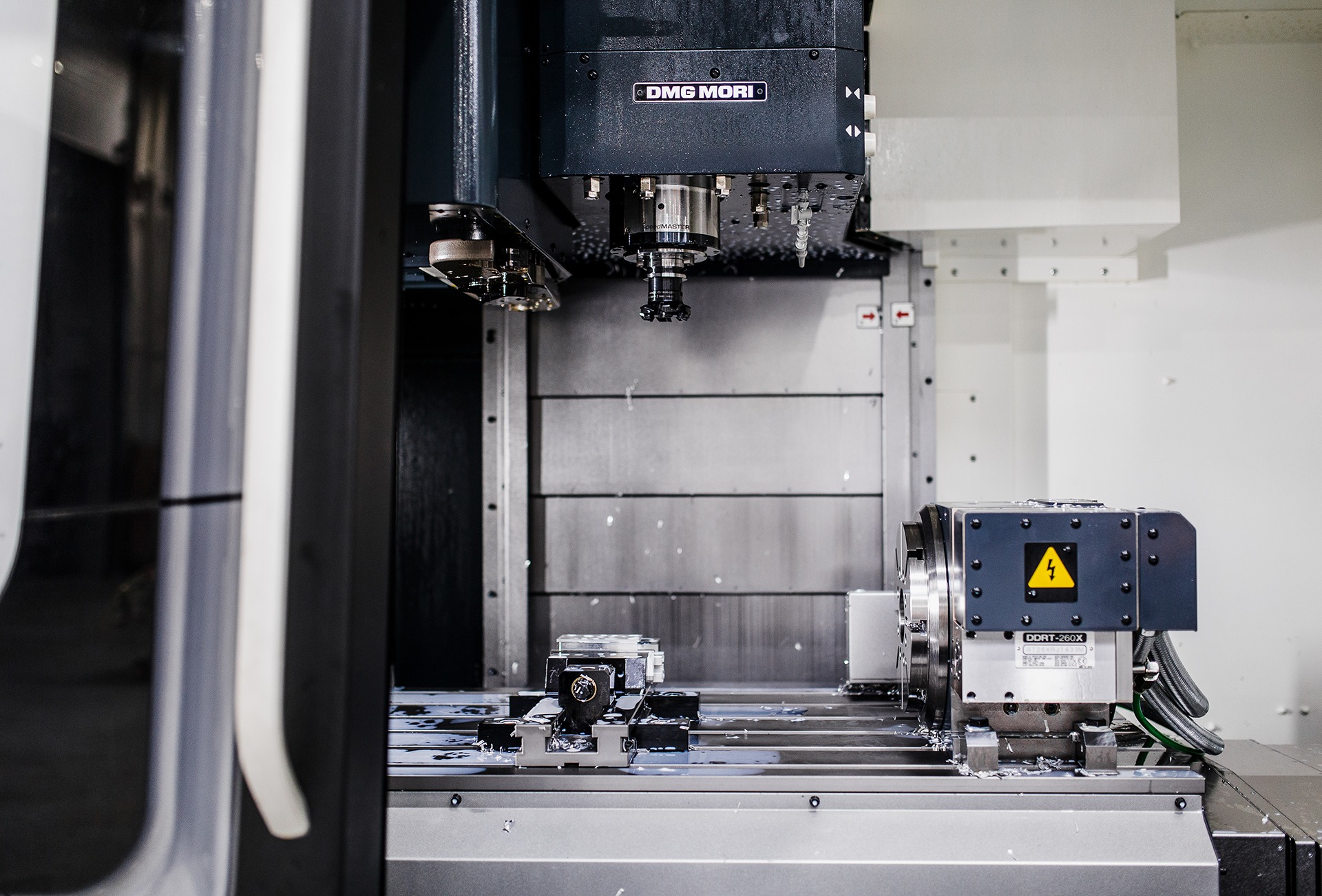 Precyzyjna obróbka metalu na frezarce CNC marki DMG MORI, widoczne chłodziwo i wióry, żółty znak ostrzegawczy na obudowie.