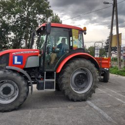OSK Tomasz Strojkowski - Czerwony traktor Zetor Proxima 90 z przyczepką i literą 'L' na boku, zaparkowany na betonowym placu z widocznym operatorem w kabinie, na tle pochmurnego nieba i drzew.