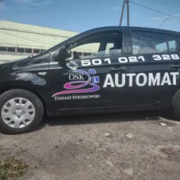 Czarny samochód z naklejkami informującymi o nauce jazdy automatem, numerem telefonu i logotypem OSK Tomasza Strojkowskiego, zaparkowany na szarym podłożu przed budynkiem.