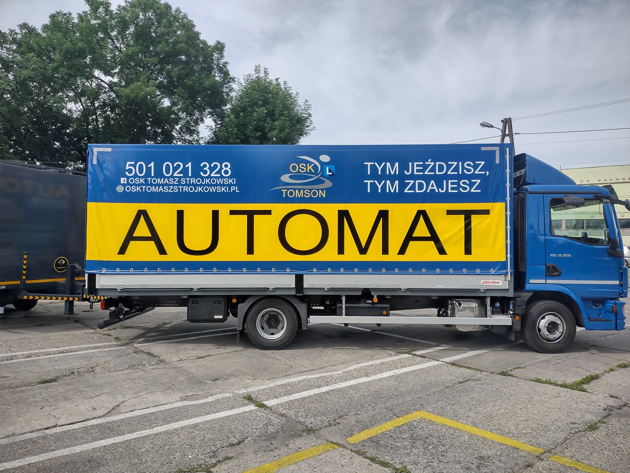 Niebieska ciężarówka z reklamą szkoły nauki jazdy z napisem 'AUTOMAT' na żółtym tle, widoczny numer telefonu i adres strony internetowej, zaparkowana na betonowym placu.