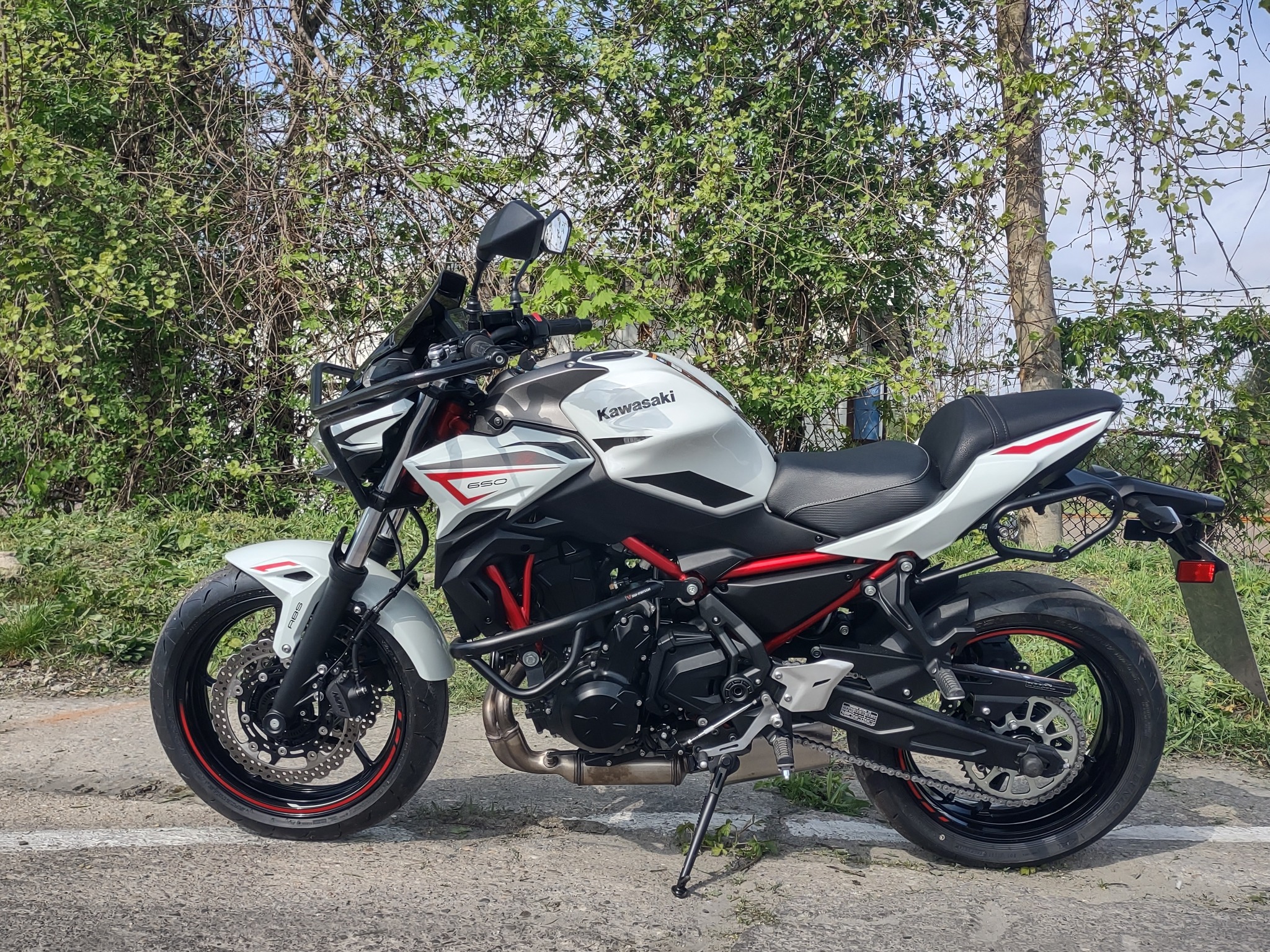 Motocykl Kawasaki Z650 w kolorze białym z czerwonymi akcentami, zaparkowany na asfalcie przed zielonymi krzewami.