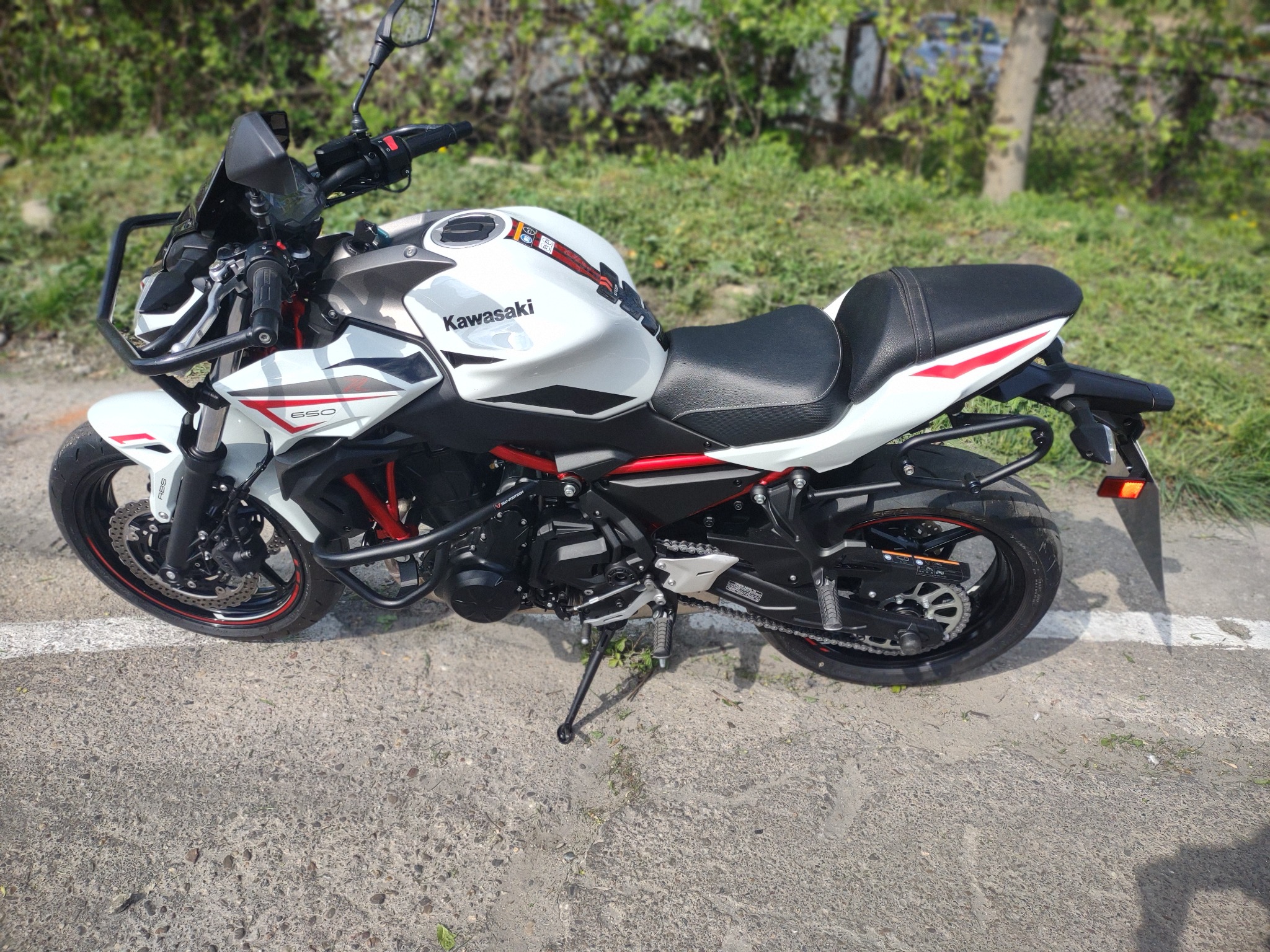 Biało-czarny motocykl Kawasaki Z650 z dodatkowym orurowaniem ochronnym przedniego reflektora, zaparkowany na szarym asfalcie w słoneczny dzień.