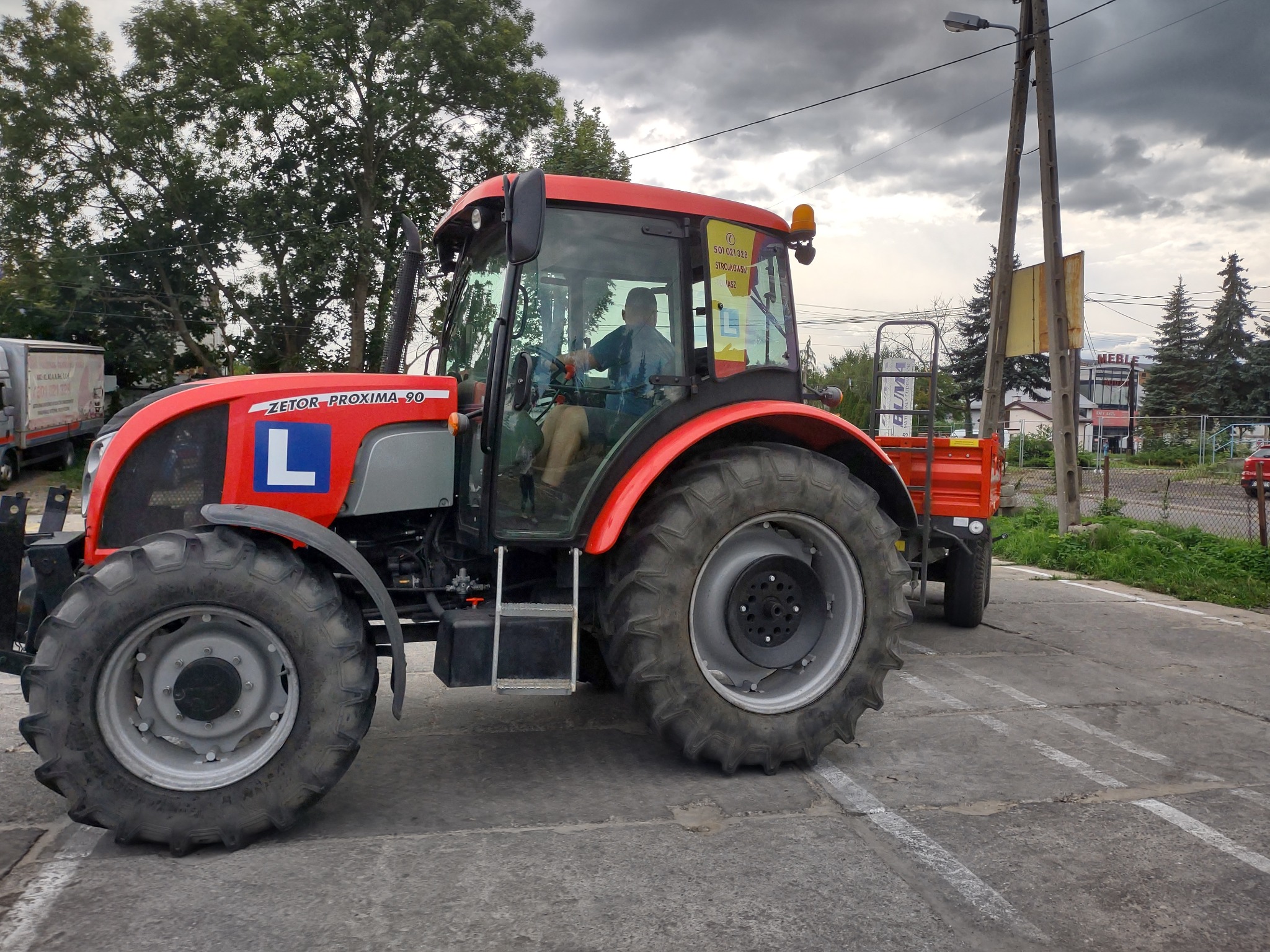 Czerwony traktor Zetor Proxima 90 z przyczepką i literą 'L' na boku, zaparkowany na betonowym placu z widocznym operatorem w kabinie, na tle pochmurnego nieba i drzew.