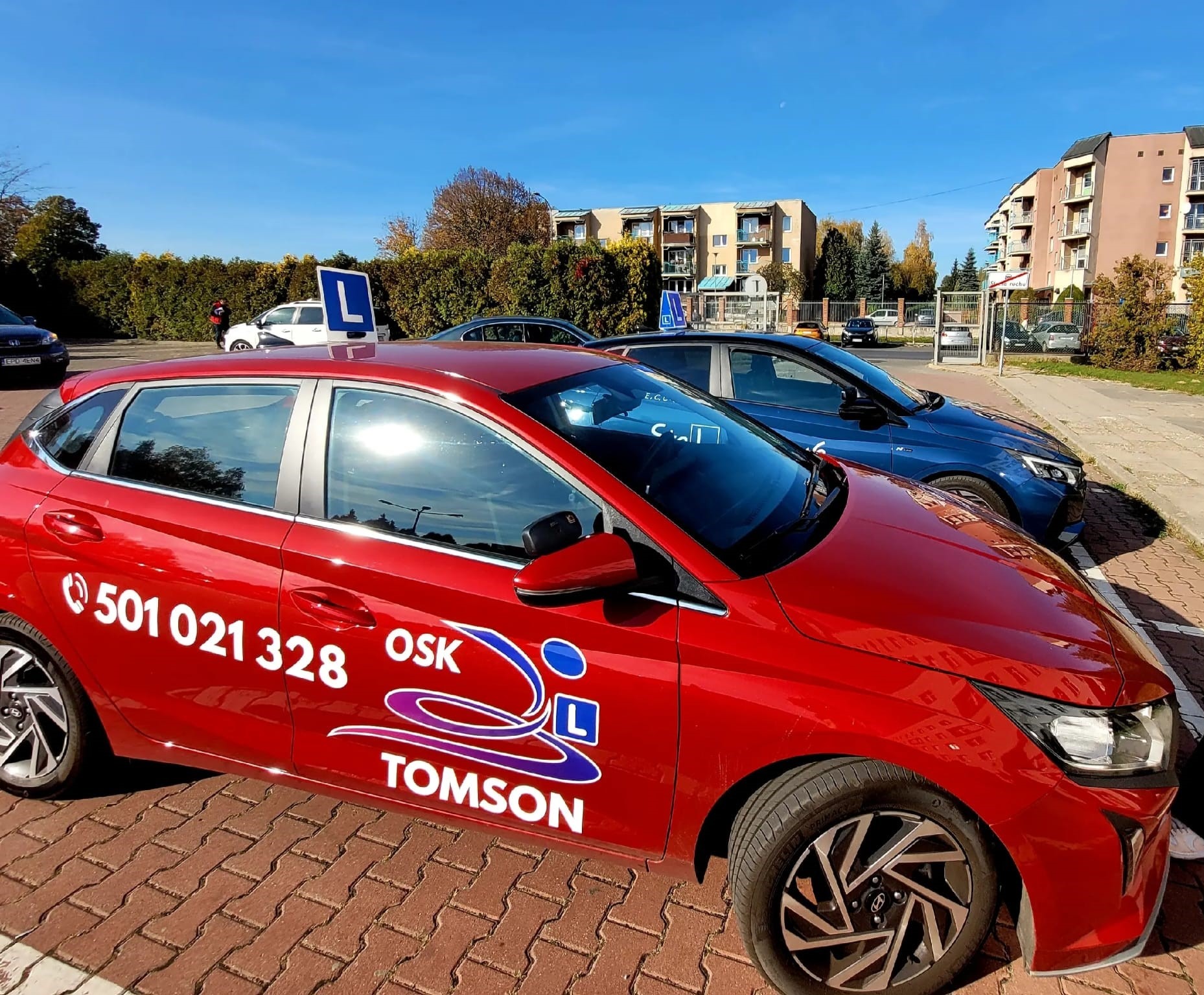 Czerwony samochód z oznaczeniami ośrodka szkolenia kierowców 'Tomson' i numerem telefonu zaparkowany na kostce brukowej, w tle budynki mieszkalne i niebieskie niebo.