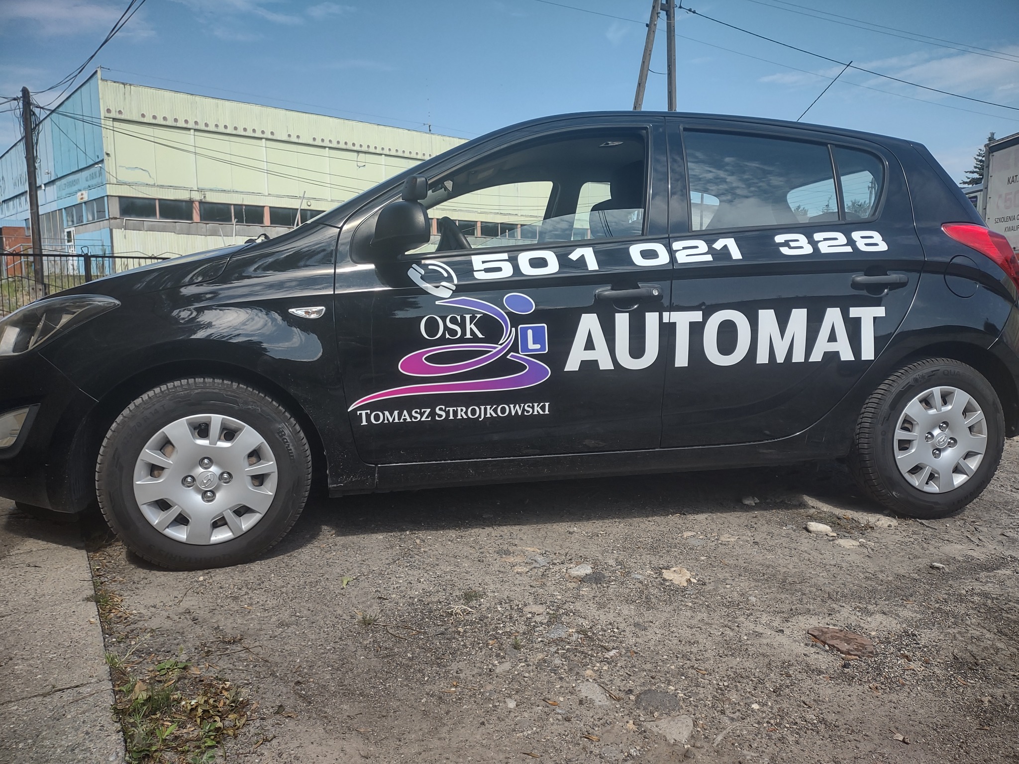 Czarny samochód z naklejkami informującymi o nauce jazdy automatem, numerem telefonu i logotypem OSK Tomasza Strojkowskiego, zaparkowany na szarym podłożu przed budynkiem.