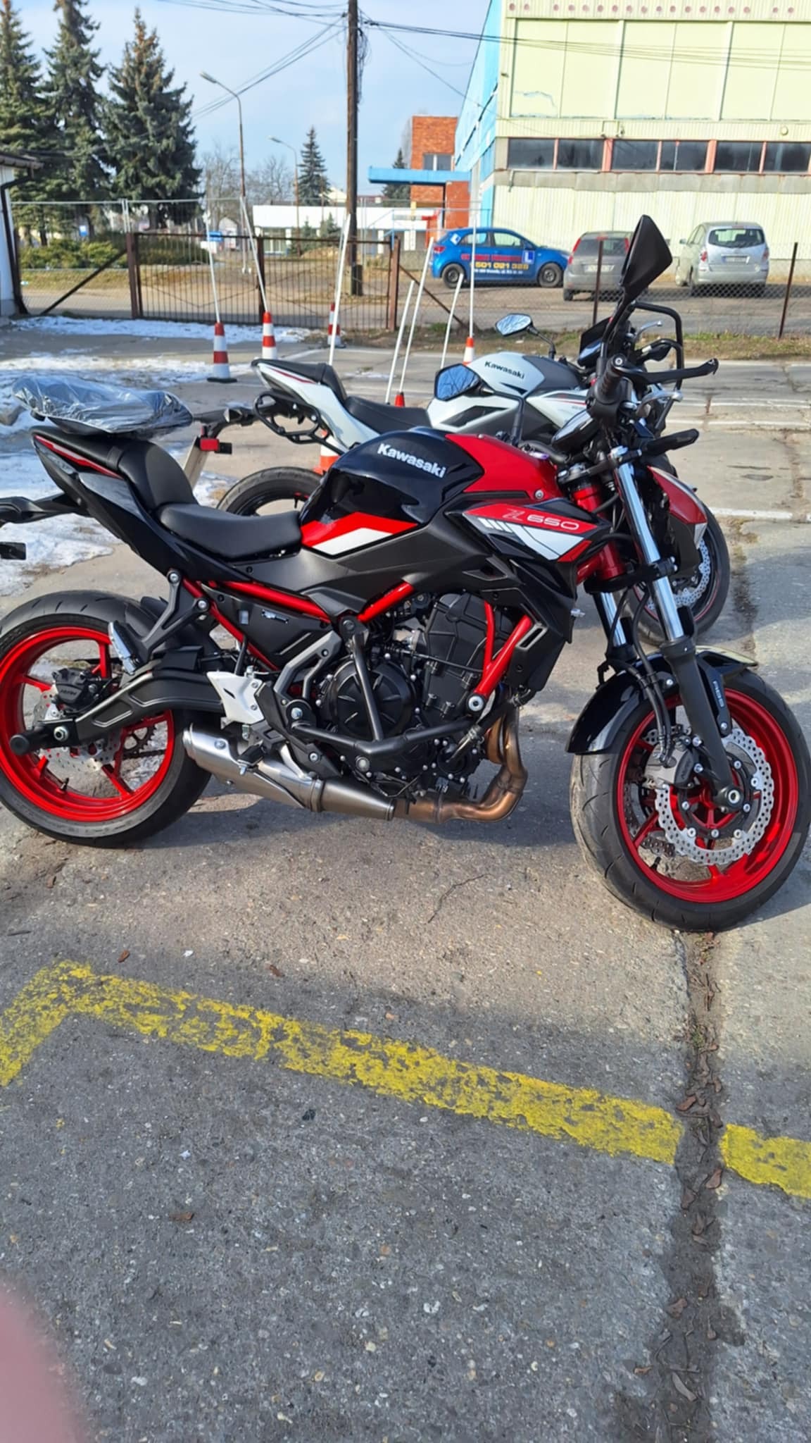 Czarno-czerwony motocykl Kawasaki Z650 zaparkowany na betonowym placu z żółtymi liniami, w tle budynek, biało-czerwone pachołki i inne motocykle.