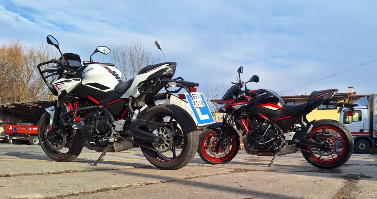 Dwa motocykle Kawasaki Z900, biały i czarny z czerwonymi akcentami, stoją obok siebie na betonowym placu; biały motocykl ma tablicę z literą 'L', sugerującą naukę jazdy.