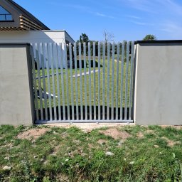 ZAPAŁA SŁAWOMIR - Nowoczesna brama wjazdowa w Strawczynie z pionowych, metalowych profili, osadzona między betonowymi słupkami. W tle trawnik i budynek z elementami drewnianymi.
