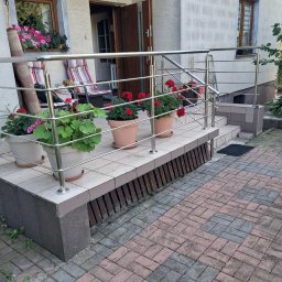 ZAPAŁA SŁAWOMIR - Nowoczesna balustrada ze stali nierdzewnej zabezpiecza schody wejściowe do domu. Donice z kwiatami zdobią stopnie. Widok na elewację i otwarte drzwi.