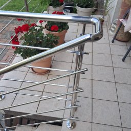 ZAPAŁA SŁAWOMIR - Błyszcząca, metalowa balustrada z poprzecznymi prętami na schodach tarasu, z doniczkami z czerwonymi kwiatami w tle, widziana z góry.
