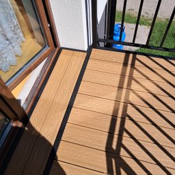 ZAPAŁA SŁAWOMIR - Nowoczesny balkon z deską kompozytową w odcieniach brązu i czarną balustradą. Widok z góry, z oknem w tle i butlą gazową widoczną przez barierki.