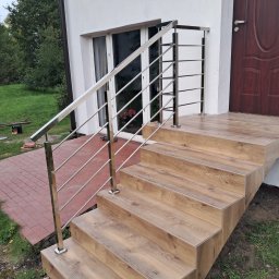 ZAPAŁA SŁAWOMIR - Zewnętrzne schody z drewnopodobnymi płytkami i balustradą ze stali nierdzewnej, prowadzące do drzwi wejściowych. Widoczne elementy wykończeniowe i detale montażowe.