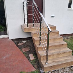 ZAPAŁA SŁAWOMIR - Schody wejściowe z drewnopodobnymi płytkami i balustradą ze stali nierdzewnej. Nowoczesne wykończenie, jasna elewacja. Widok z dołu, detale poręczy i stopnic.