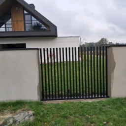 Nowoczesna, czarna brama wjazdowa z pionowych profili, osadzona w betonowym murze. W tle dom z drewnianymi elementami i trójkątnym oknem na poddaszu.