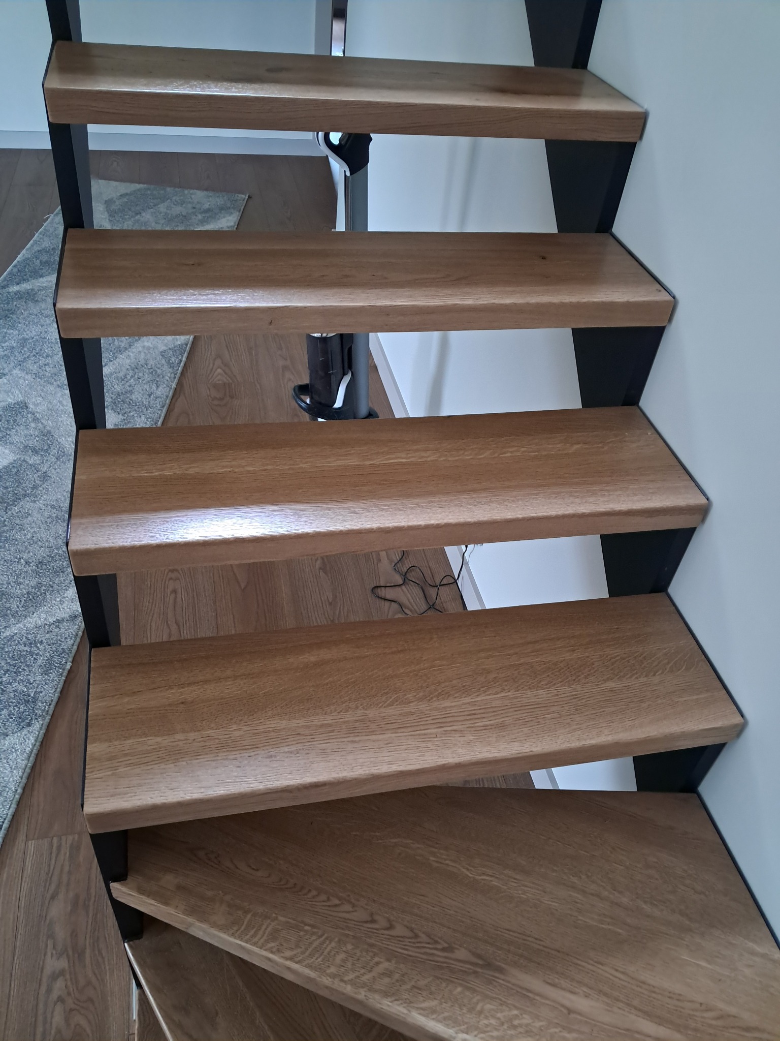 Drewniane stopnie schodów na czarnej metalowej konstrukcji, widok z góry, zarys dywanu i odkurzacza w tle.