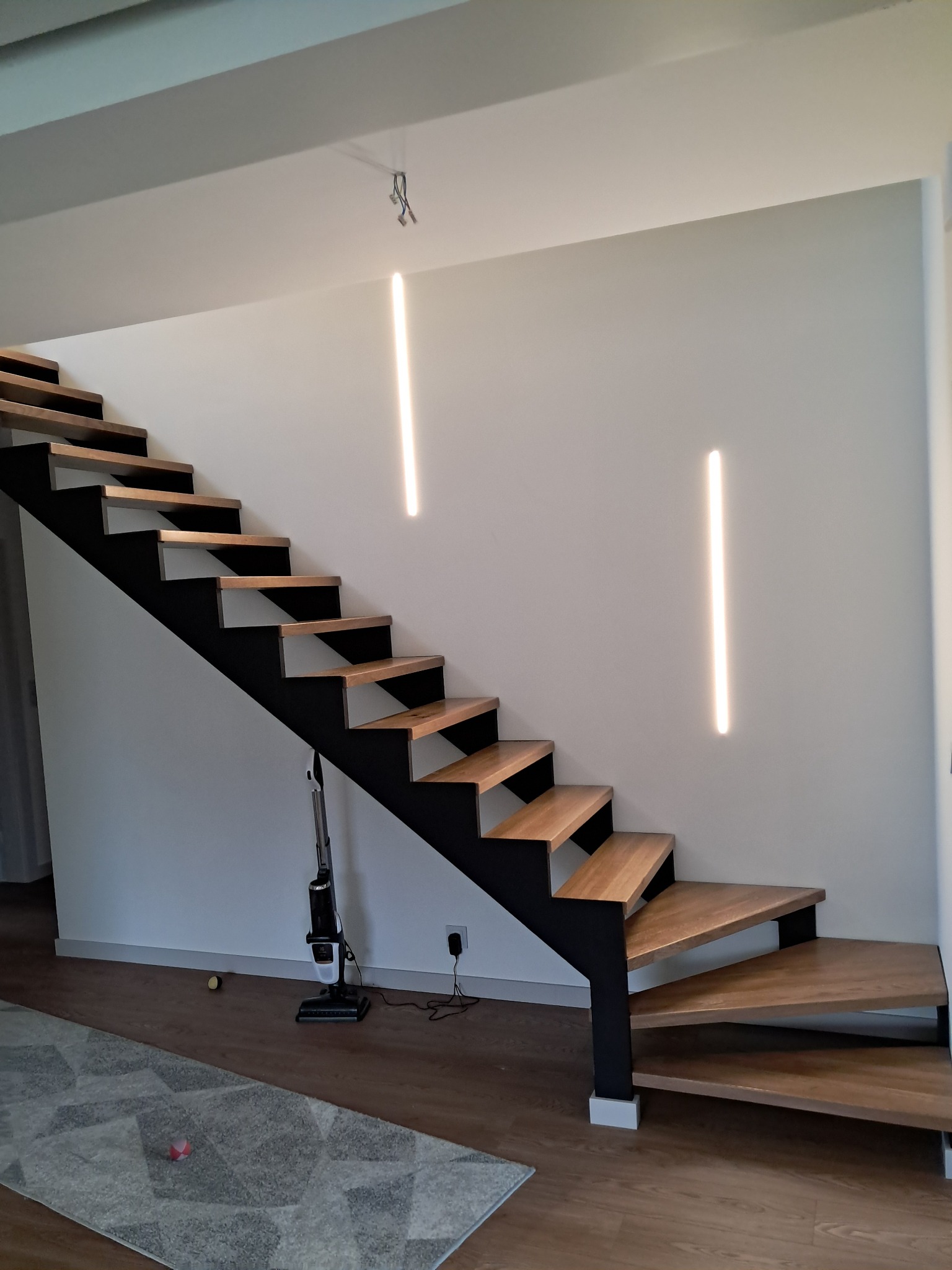 Nowoczesne schody metalowe z drewnianymi stopniami, oświetlone pionowymi lampami LED na ścianie, widoczne z boku, z odkurzaczem bezprzewodowym i dywanem w tle.
