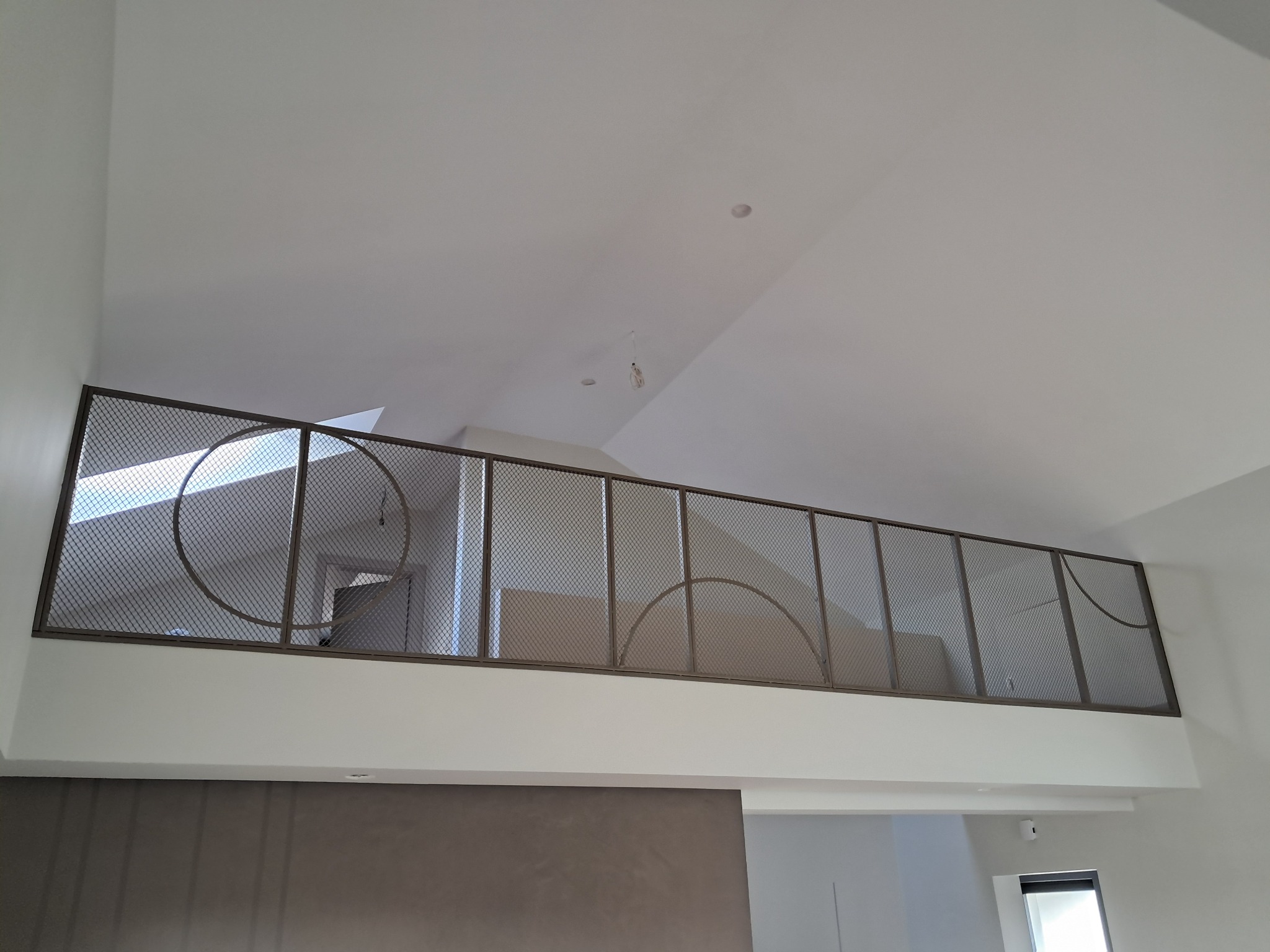 Metalowa balustrada z siatki z dekoracyjnymi elementami w kształcie okręgów, zamontowana na antresoli w nowoczesnym wnętrzu z białymi ścianami i skośnym sufitem.