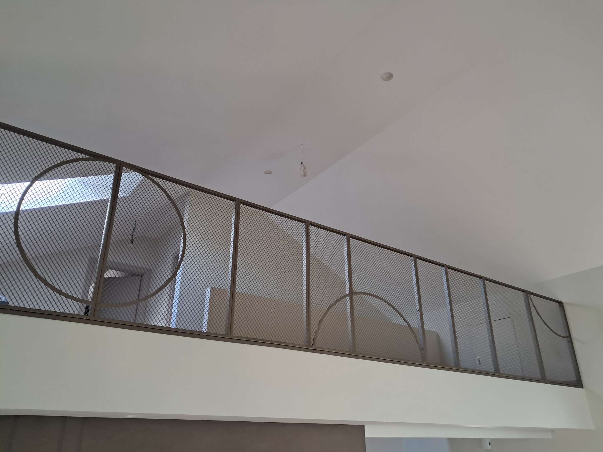 Metalowa balustrada z siatki z dekoracyjnymi elementami w kształcie półokręgów i okręgów, widok z dołu na jasnym tle ściany i sufitu.