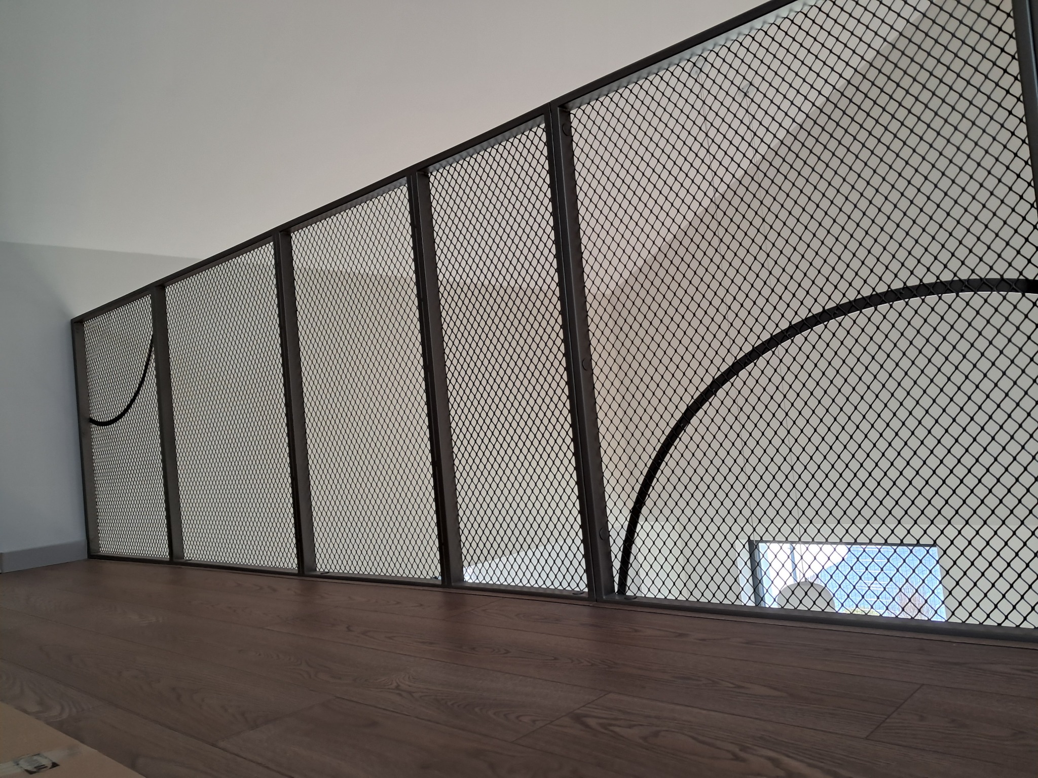 Metalowa siatka zabezpieczająca schody na antresoli z drewnianą podłogą i widokiem na okno zza niej.