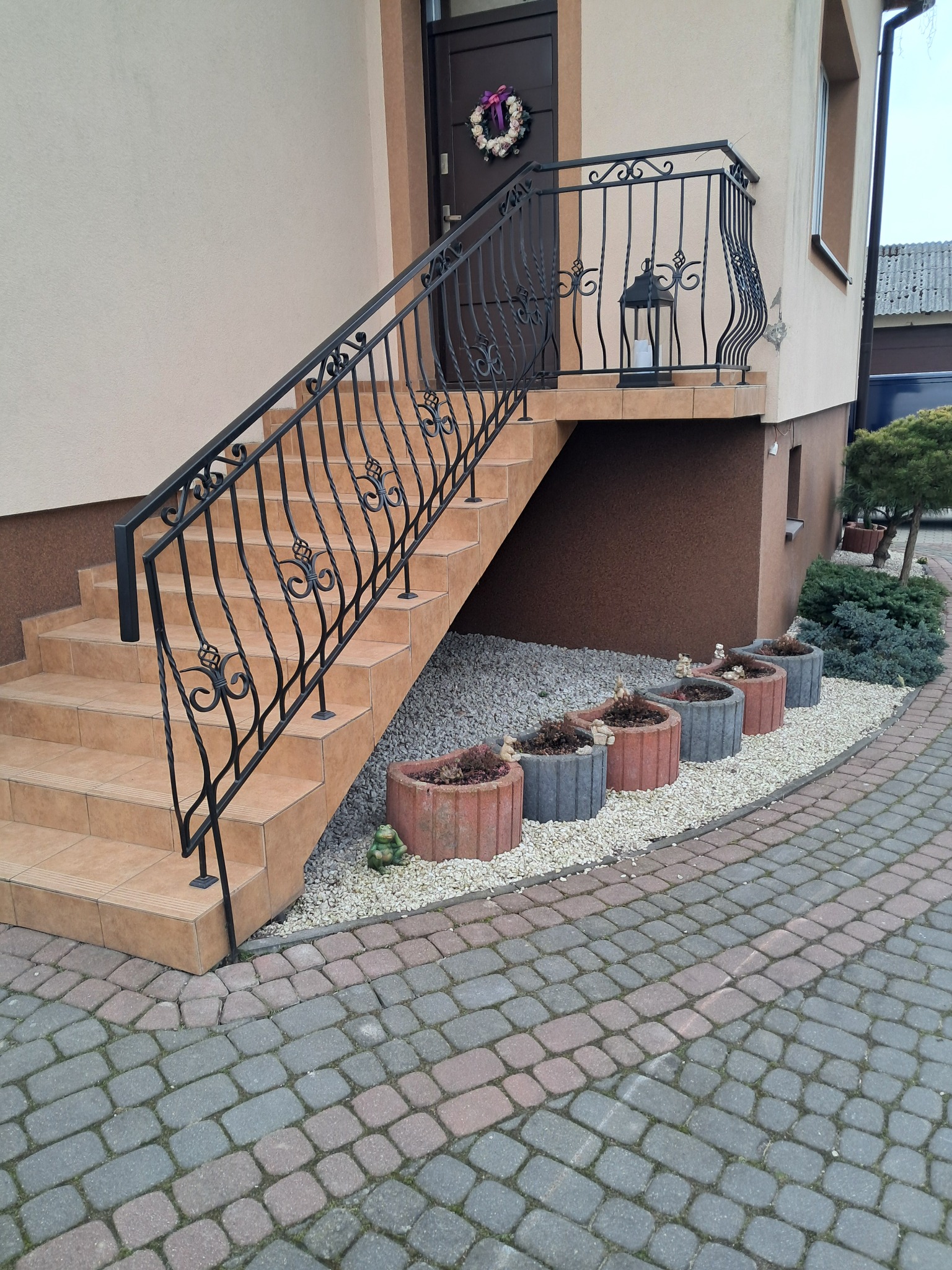 Kute, czarne balustrady schodowe z dekoracyjnymi elementami na zewnątrz budynku, z widoczną latarnią na podeście i ozdobnym wieńcem na drzwiach.