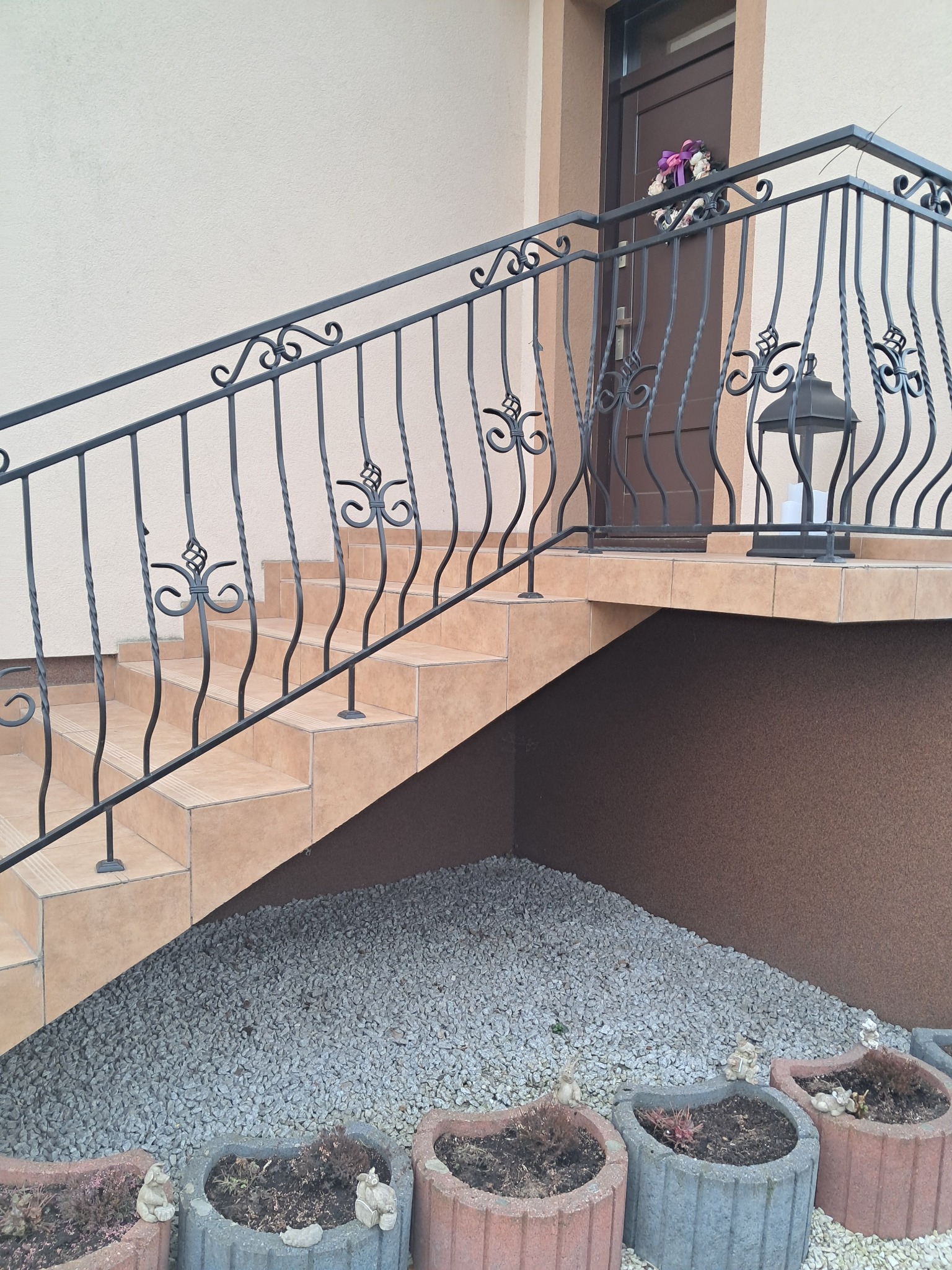 Zewnętrzne schody z beżowych płytek prowadzą do brązowych drzwi, zabezpieczone czarną, kutą balustradą z dekoracyjnymi elementami. Pod schodami ozdobne kamienie i donice.