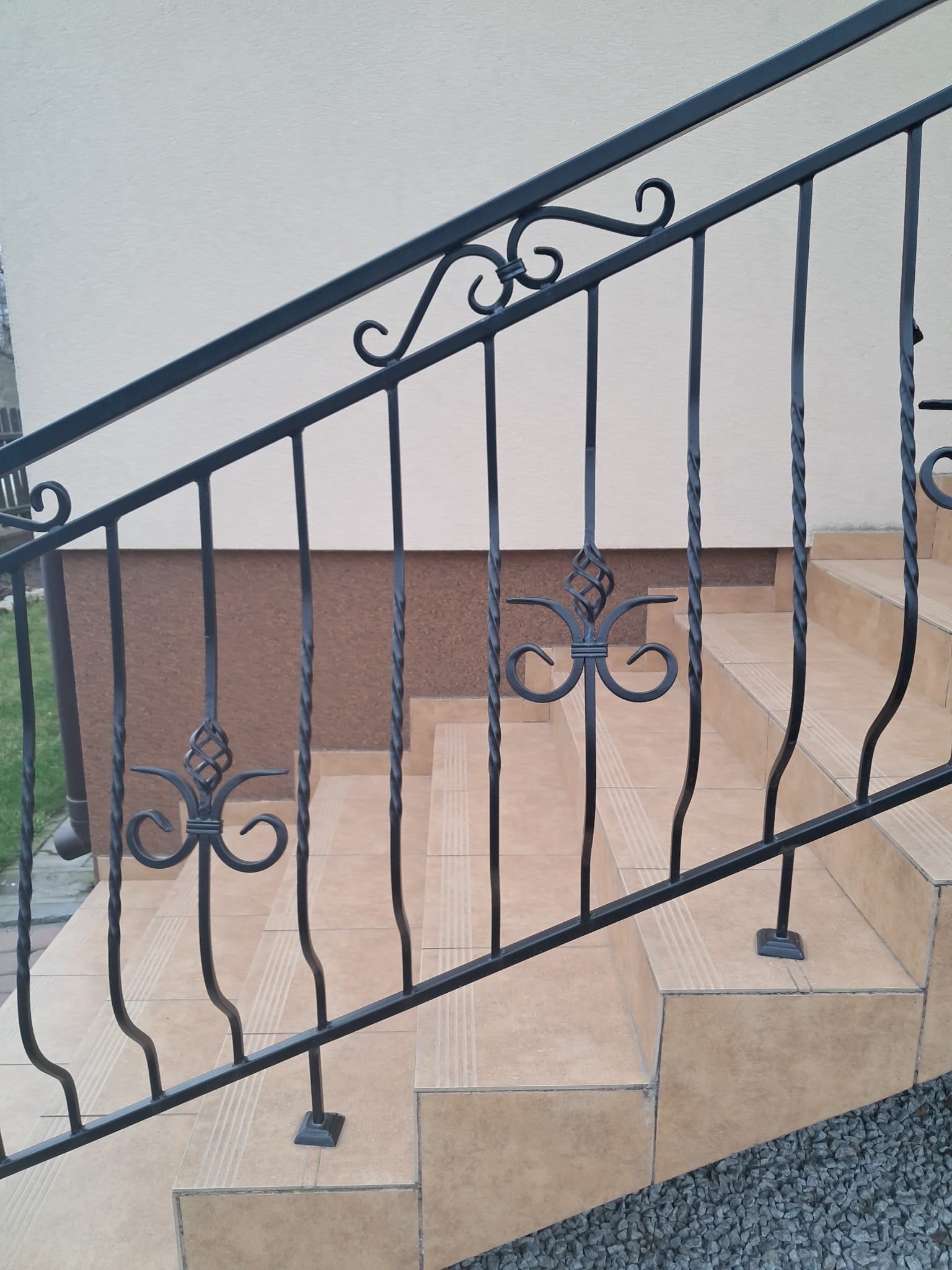 Metalowa balustrada schodowa z dekoracyjnymi elementami w kształcie ślimaków i skręconych prętów, zamontowana na beżowych, wyłożonych płytkami schodach zewnętrznych.