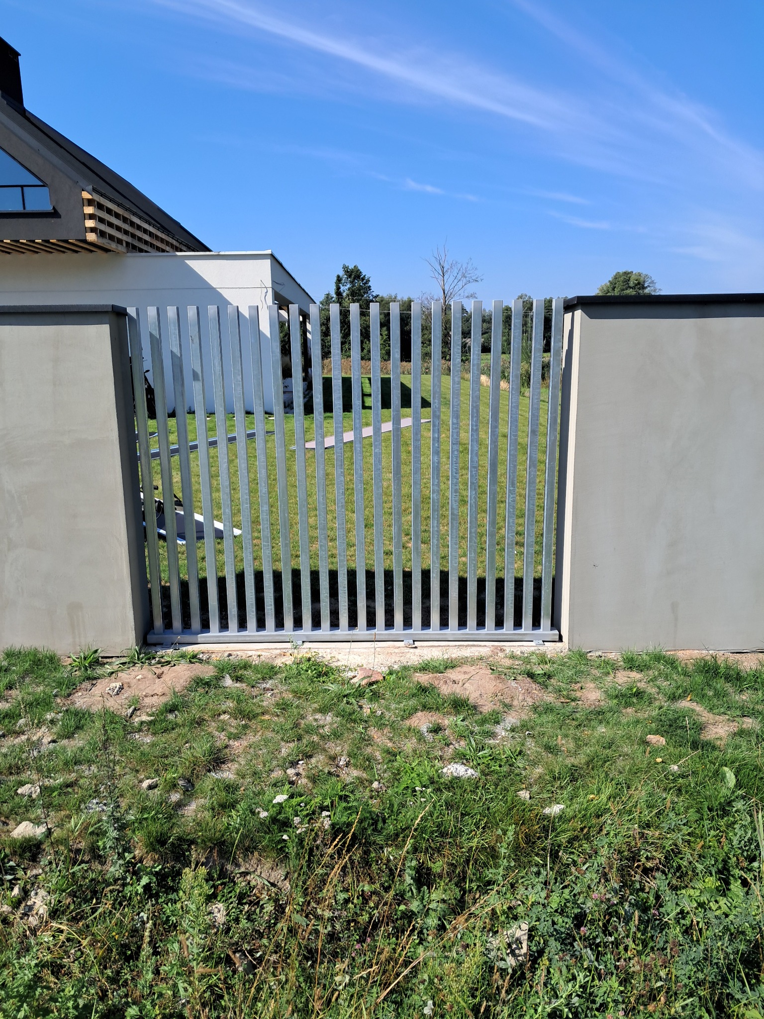 Nowoczesna brama wjazdowa w Strawczynie z pionowych, metalowych profili, osadzona między betonowymi słupkami. W tle trawnik i budynek z elementami drewnianymi.