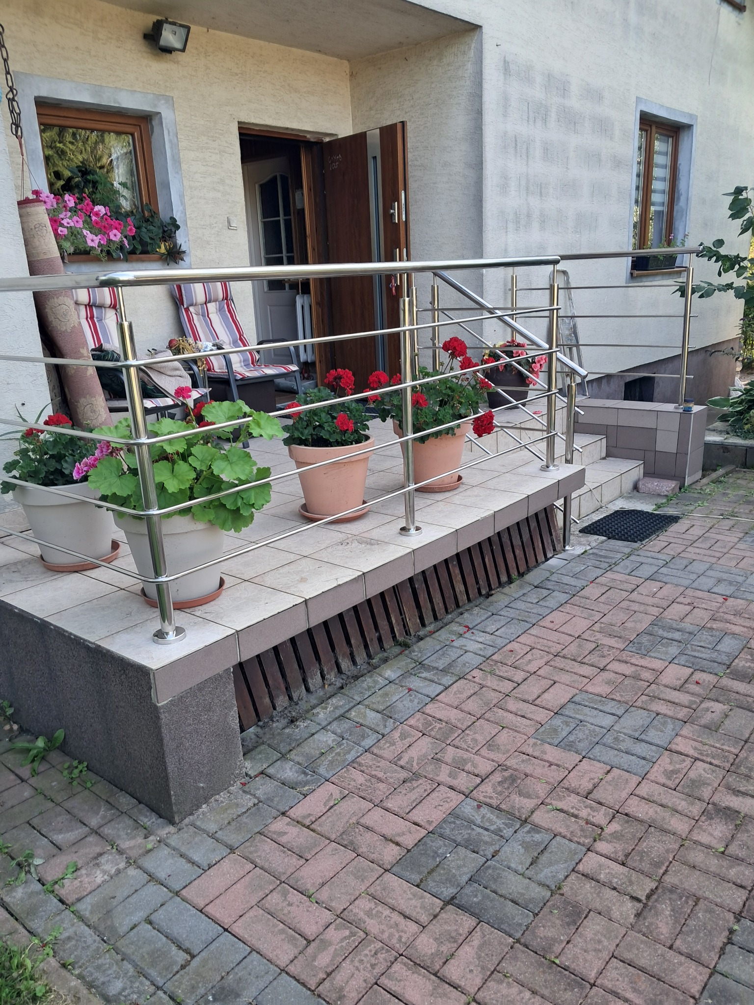Nowoczesna balustrada ze stali nierdzewnej zabezpiecza schody wejściowe do domu. Donice z kwiatami zdobią stopnie. Widok na elewację i otwarte drzwi.