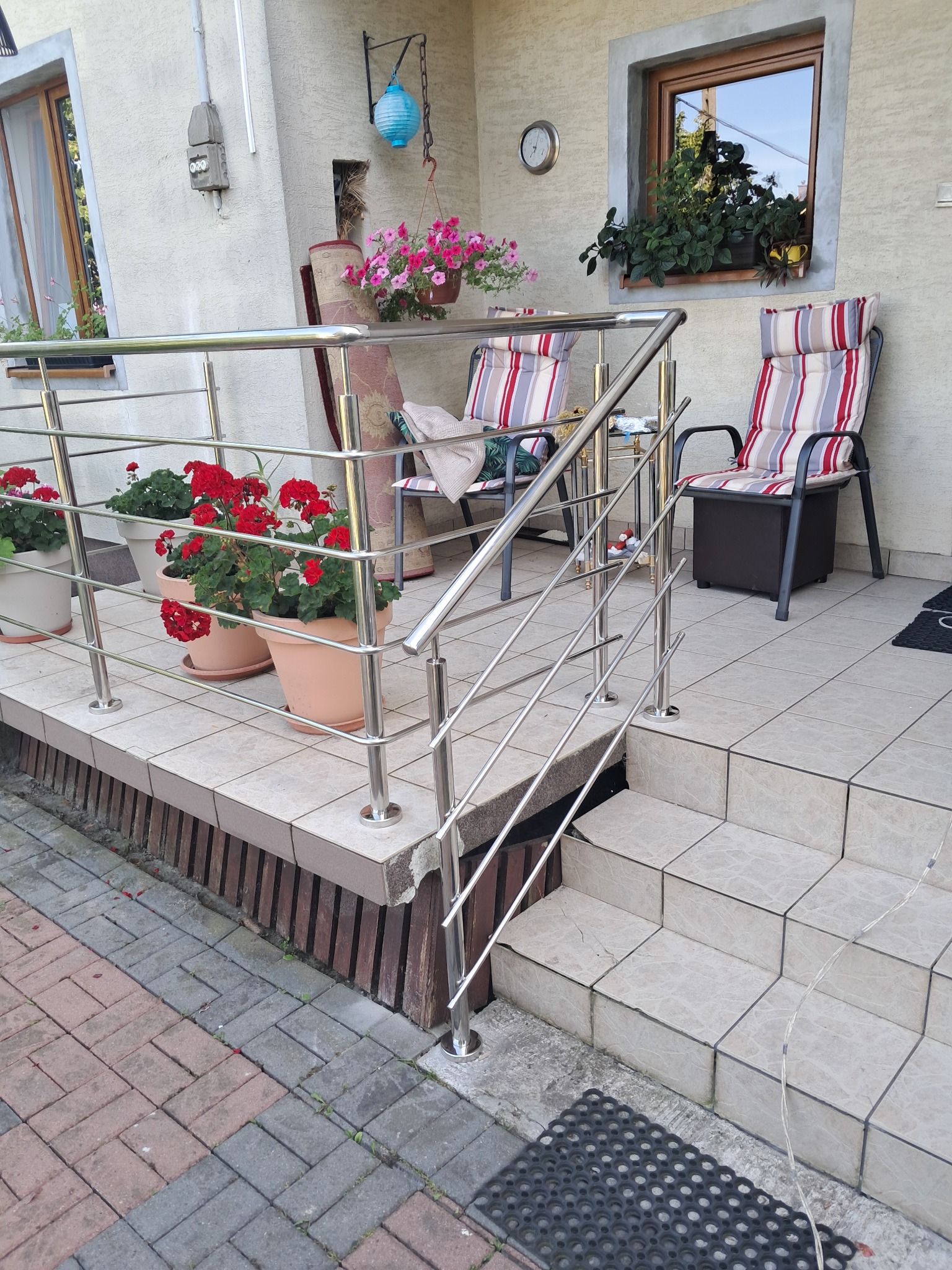 Błyszcząca, metalowa balustrada z poręczą na schodach wejściowych do domu, z kwiatami w donicach i dwoma krzesłami w tle. Ujęcie z dołu, jasne światło dzienne.