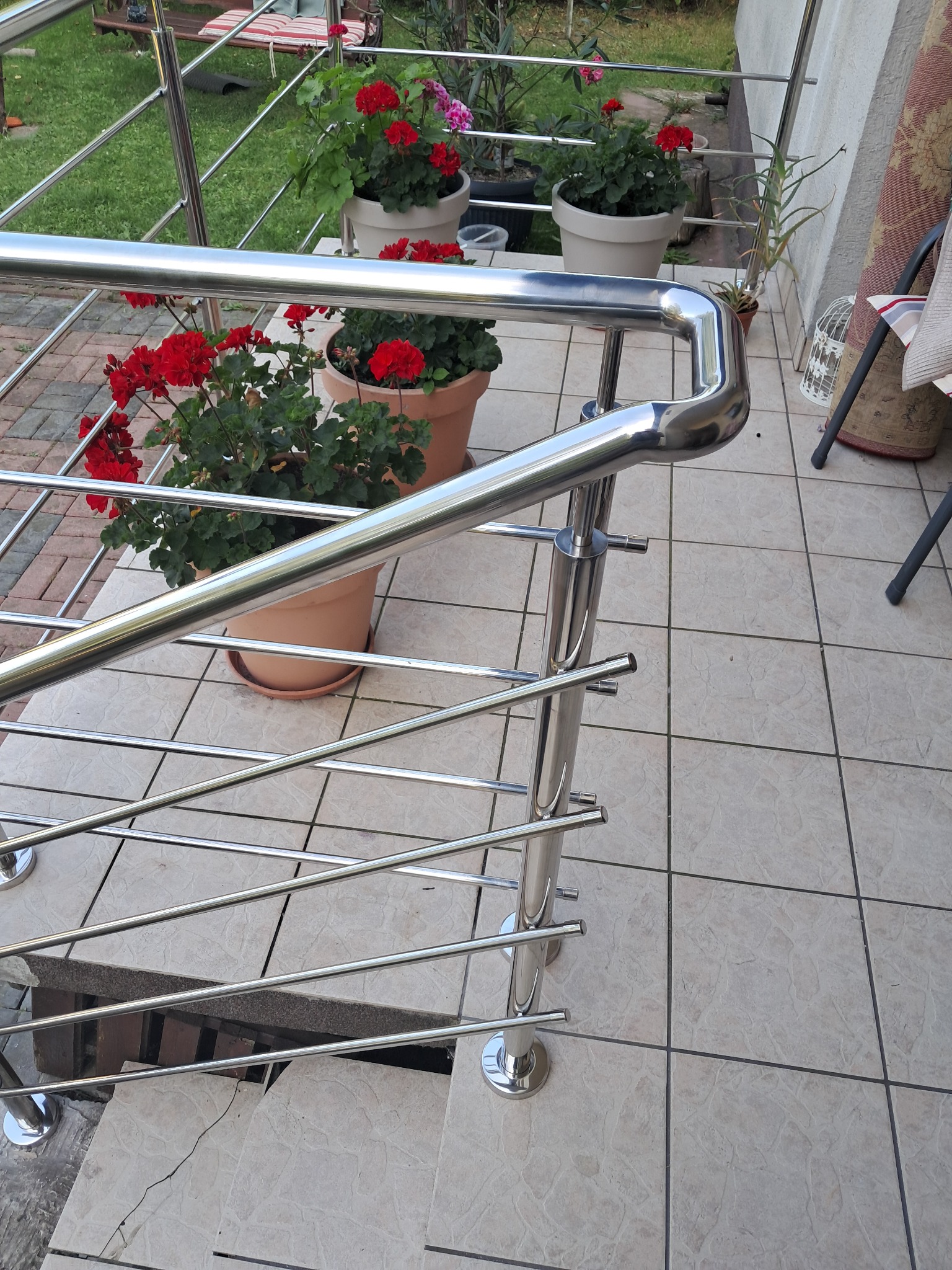 Błyszcząca, metalowa balustrada z poprzecznymi prętami na schodach tarasu, z doniczkami z czerwonymi kwiatami w tle, widziana z góry.
