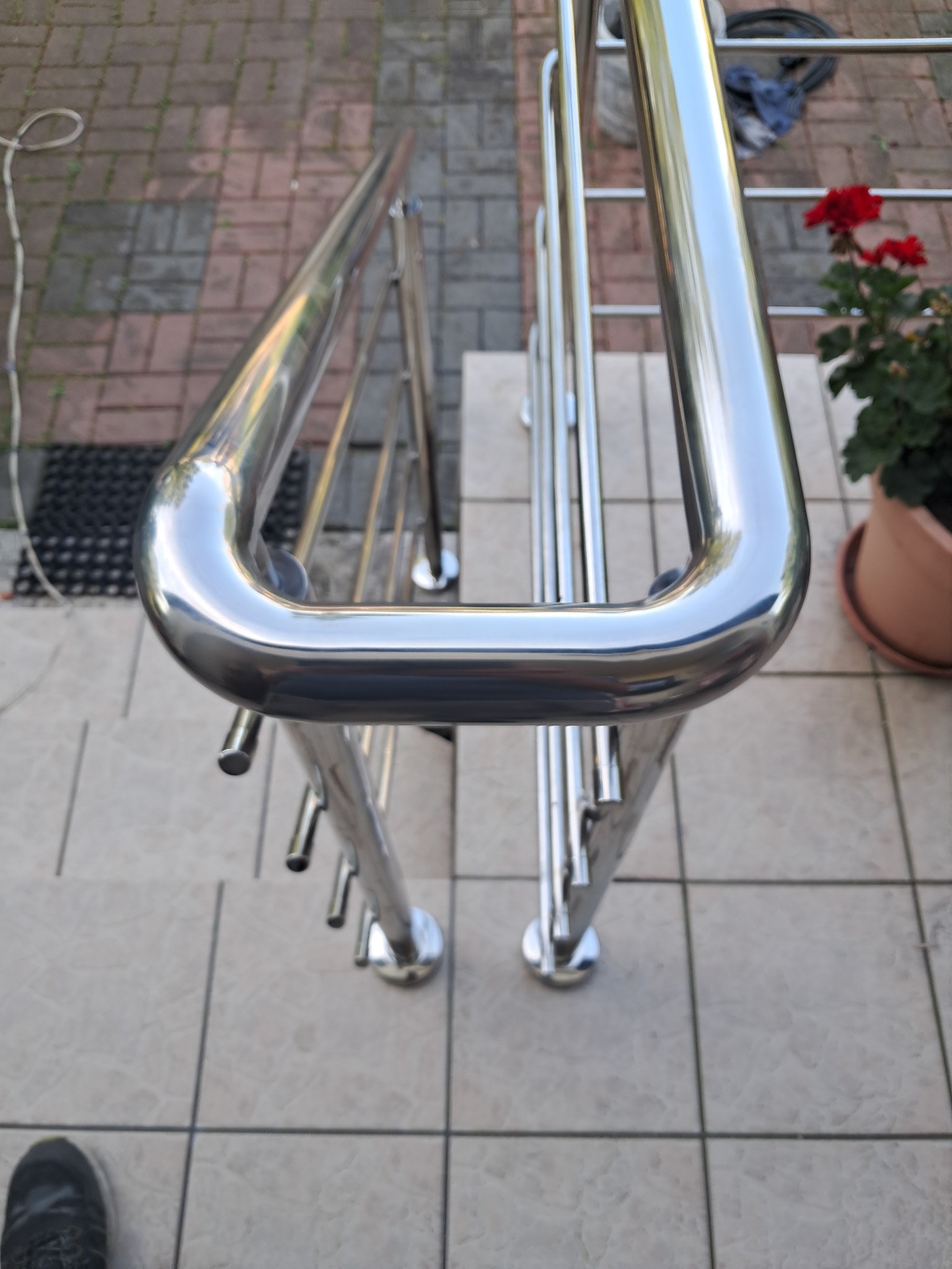 Polerowana balustrada nierdzewna na zewnątrz budynku, z kwiatami w doniczce w tle. Widok z góry, podkreślający detale wykończenia.