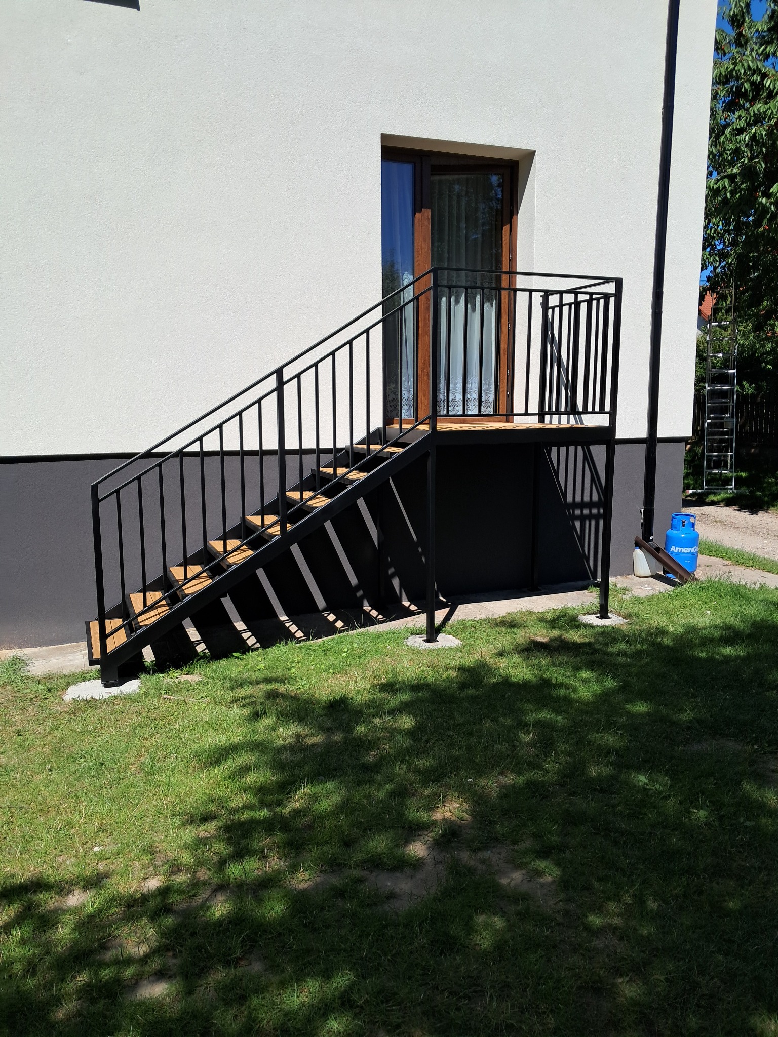 Zewnętrzne schody metalowe z drewnianymi stopniami prowadzące do drzwi. Czarna balustrada. Butla gazowa i drabina w tle.