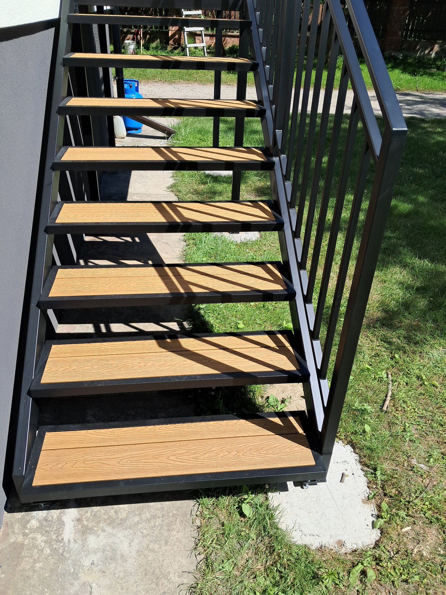 Zewnętrzne schody metalowe z drewnopodobnymi stopniami i czarną balustradą, prowadzące do góry, z widoczną zieloną trawą w tle.