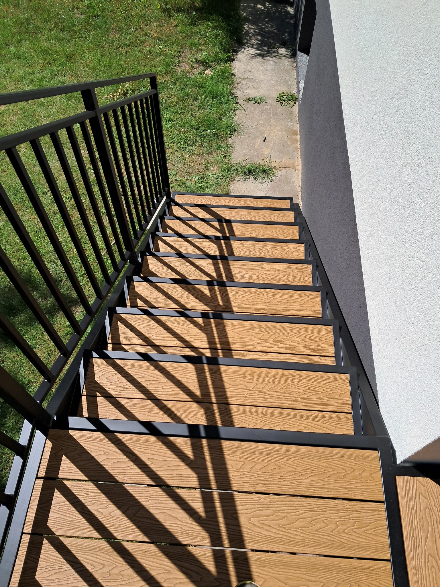 Schody zewnętrzne z metalową balustradą i drewnopodobnymi stopniami w jasnym otoczeniu. Widok z góry podkreśla geometrię konstrukcji i cień balustrady.