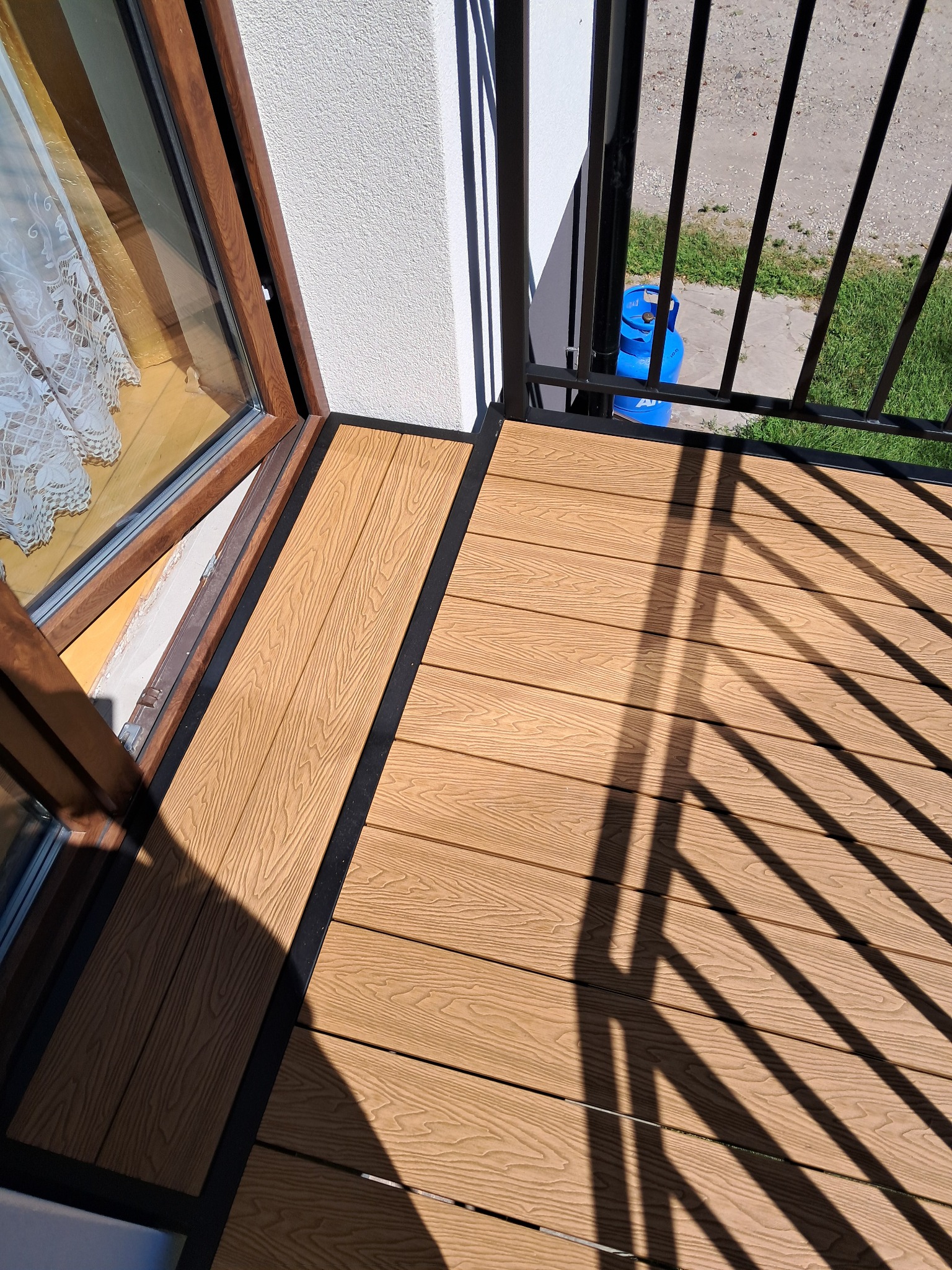Nowoczesny balkon z deską kompozytową w odcieniach brązu i czarną balustradą. Widok z góry, z oknem w tle i butlą gazową widoczną przez barierki.