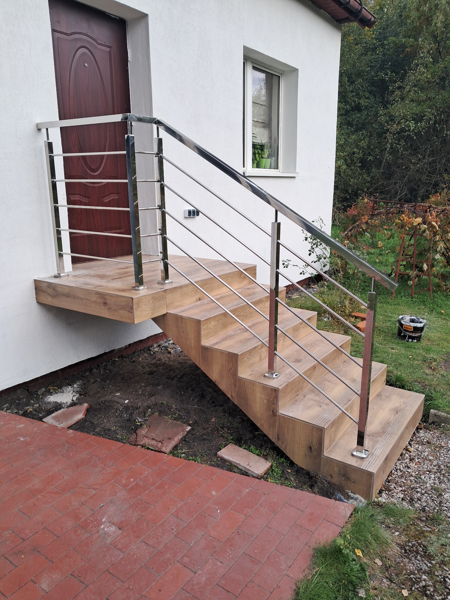 Drewniane schody zewnętrzne z nierdzewną balustradą, prowadzące do drzwi wejściowych. Widoczne płytki na stopniach i nowoczesny design balustrady.