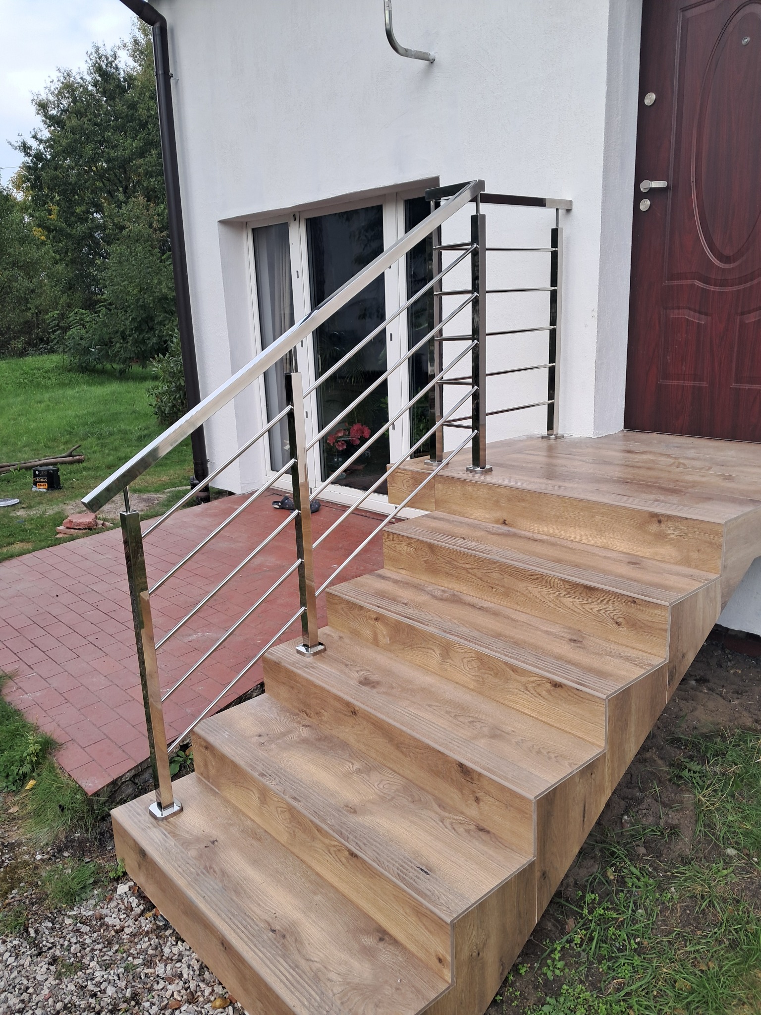 Zewnętrzne schody z drewnopodobnymi płytkami i balustradą ze stali nierdzewnej, prowadzące do drzwi wejściowych. Widoczne elementy wykończeniowe i detale montażowe.