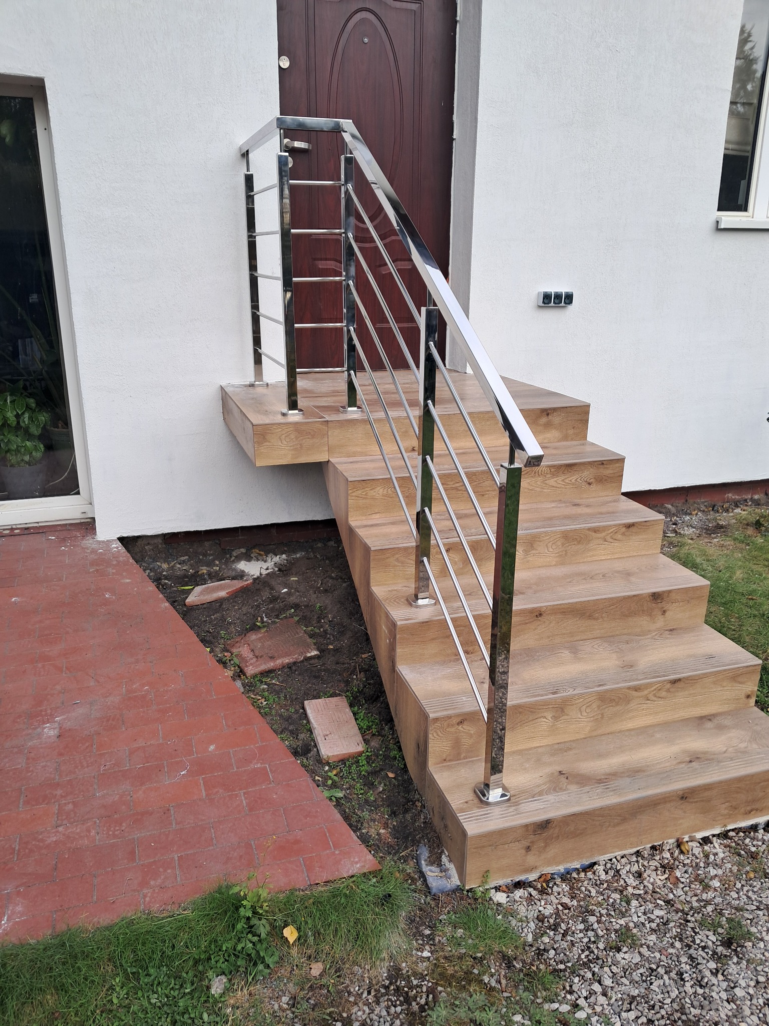 Schody wejściowe z drewnopodobnymi płytkami i balustradą ze stali nierdzewnej. Nowoczesne wykończenie, jasna elewacja. Widok z dołu, detale poręczy i stopnic.