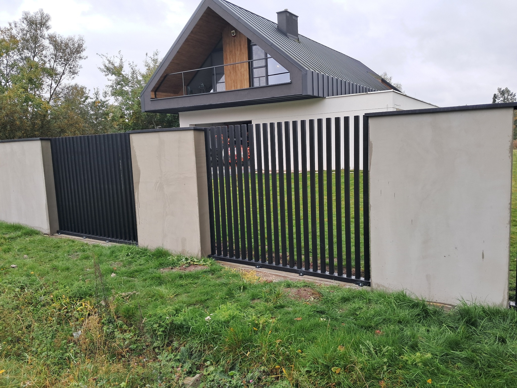 Nowoczesne ogrodzenie panelowe w kolorze czarnym, z betonowymi słupkami. W tle widoczny dom z charakterystycznym dachem. Całość otoczona zielonym trawnikiem.