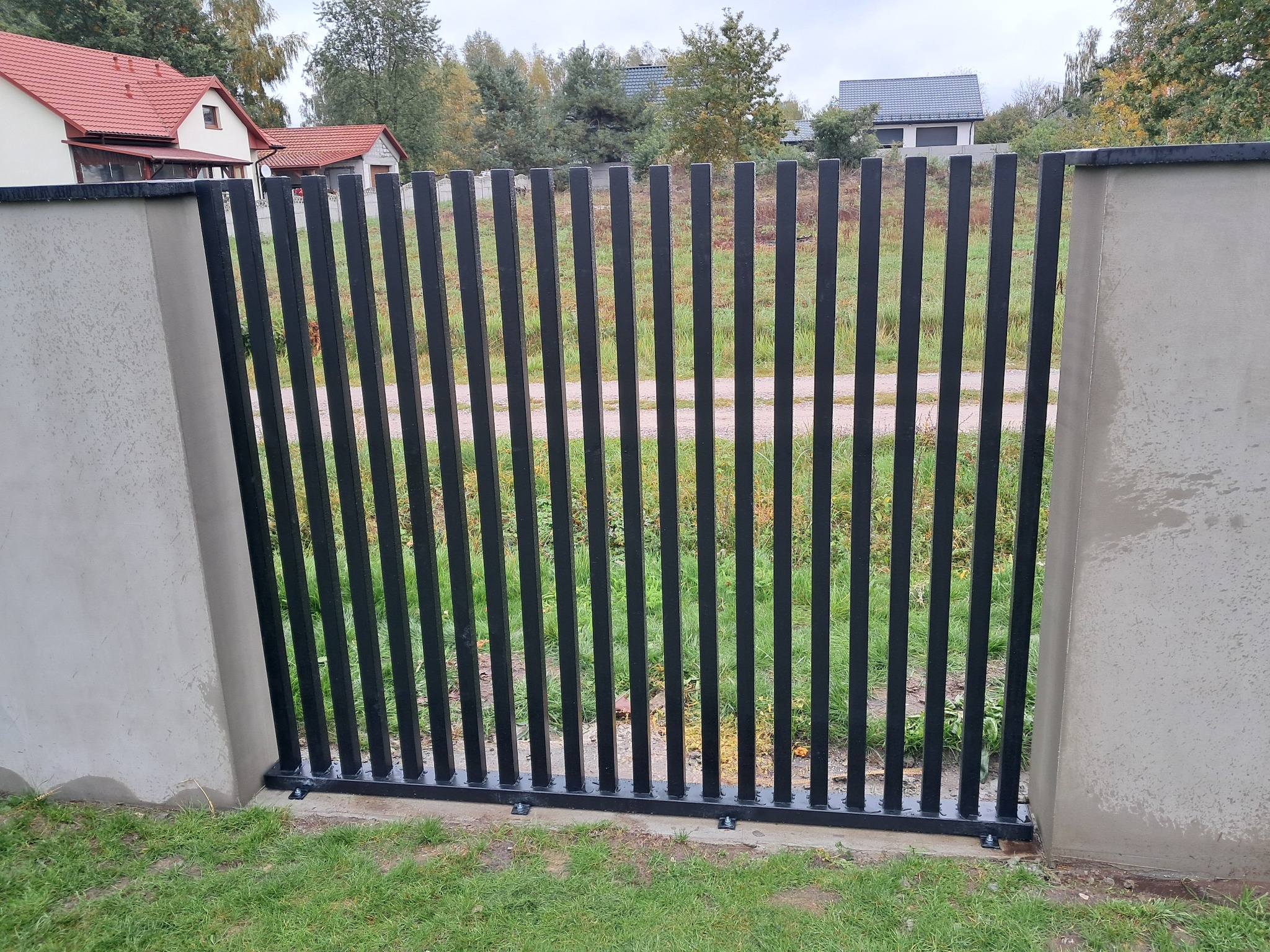 Nowoczesna, czarna brama wjazdowa z pionowych profili, osadzona między betonowymi słupkami na tle zieleni. Widoczne domy w oddali.
