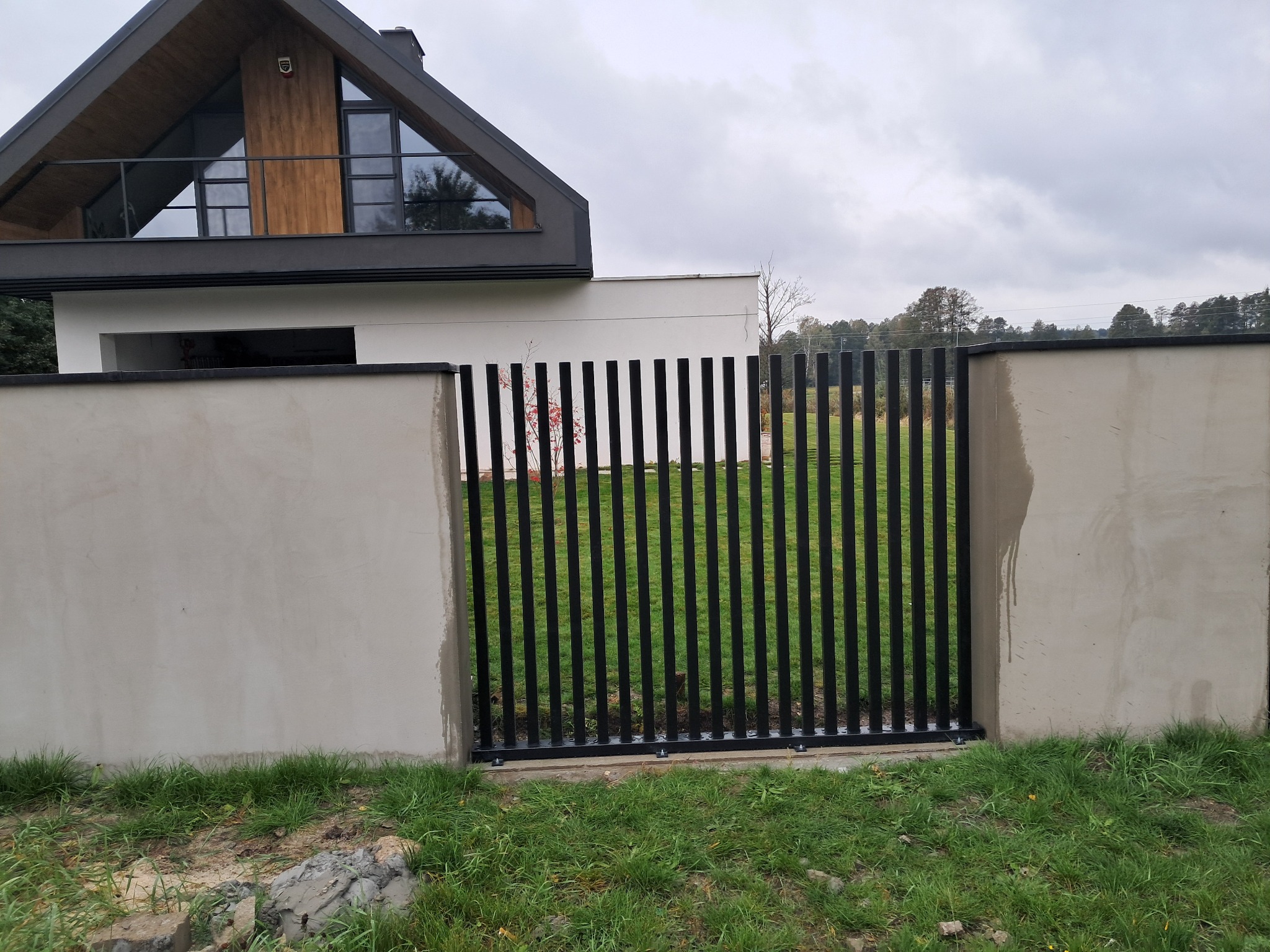 Nowoczesna, czarna brama wjazdowa z pionowych profili, osadzona w betonowym murze. W tle dom z drewnianymi elementami i trójkątnym oknem na poddaszu.