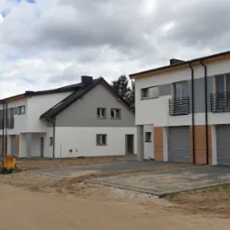 Szereg nowych, dwukondygnacyjnych domów z garażami w stanie deweloperskim, z szarymi dachami i elewacjami w odcieniach bieli i szarości, na niezagospodarowanym terenie.