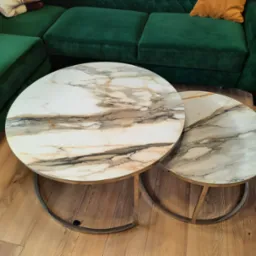 Dwa okrągłe stoliki kawowe z blatami imitującymi marmur, ustawione jeden obok drugiego na drewnianej podłodze, z fragmentem zielonej kanapy w tle.