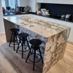 Wyspa kuchenna z blatem z kamienia o unikalnym wzorze przypominającym mapę, otoczona trzema czarnymi hokerami na drewnianej podłodze, z widocznym oknem i elementami wyposażenia kuchni w tle.