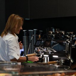 Tryb Manualny by Jakub Szołdra-Laszczyk - Kobieta w białej koszuli obsługuje profesjonalny ekspres do kawy, przygotowując napój. W tle widoczne kubki i filiżanki.