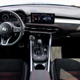 Tryb Manualny by Jakub Szołdra-Laszczyk - Wnętrze samochodu Alfa Romeo z czarną deską rozdzielczą obszytą czerwoną nicią, widoczny ekran nawigacji prezentujący model samochodu, kierownica z logo marki oraz automatyczna skrzynia biegów.