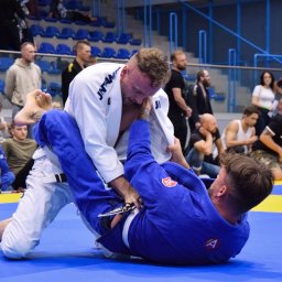 Tryb Manualny by Jakub Szołdra-Laszczyk - Dwóch mężczyzn w kimonach, białe i niebieskie, walczących na niebiesko-żółtej macie podczas zawodów brazylijskiego jiu-jitsu, z publicznością w tle.