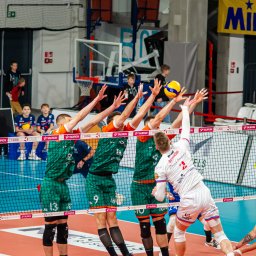 Tryb Manualny by Jakub Szołdra-Laszczyk - Mecz siatkówki: blok czterech zawodników w zielono-pomarańczowych strojach próbujących zatrzymać atakującego w białym stroju z numerem 2, w tle widownia i banery reklamowe.